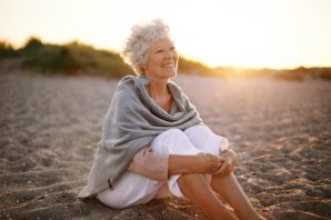 Du siehst eine gesunde Dame mittleren Alters gemütlich und entspannt am Strand im Sonnenuntergang sitzen.